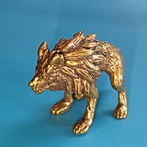 Puede incluir: Pequeña y detallada figura de lobo de latón. La escultura está en posición de caminar, con pelaje texturizado y una cara detallada. El lobo es de color dorado, sobre un fondo azul.