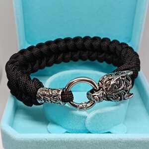 Op de afbeelding: Een zwart gevlochten armband met zilverkleurige metalen details. De armband heeft een wolfskopsluiting en decoratieve eindstukken. De armband wordt gepresenteerd in een lichtblauwe sieradendoos.