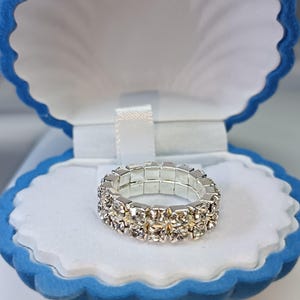 Puede incluir: Un anillo plateado con dos filas de brillantes pedrería transparente. El anillo se exhibe dentro de un joyero azul en forma de concha con interior blanco y una cinta blanca.