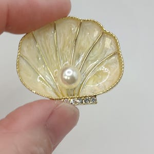 Op de afbeelding: Schelpvormige broche met een crèmekleurige emaille afwerking en een goudkleurige rand. De broche heeft een centrale parel en een rij kleine, heldere kristallen. Het ontwerp doet denken aan oceaanthema's.