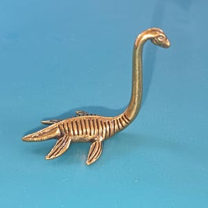 Peut inclure: Figurine de dinosaure de couleur laiton, avec un long cou et quatre appendices en forme de nageoires. Le dinosaure a un corps texturé et détaillé, sur fond bleu. La figurine est un objet décoratif, peut-être une broche ou une petite sculpture.