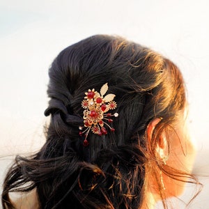 Peut inclure: Une barrette dor&eacute;e orn&eacute;e de pierres pr&eacute;cieuses rouges et transparentes et d'&eacute;l&eacute;ments floraux est fix&eacute;e dans des cheveux fonc&eacute;s. Cet accessoire de cheveux d&eacute;coratif est probablement destin&eacute; &agrave; une occasion sp&eacute;ciale, sur un fond flou.