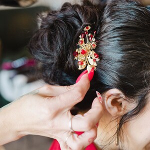 Peut inclure: Un accessoire pour cheveux dor&eacute; avec des pierres pr&eacute;cieuses rouges et des d&eacute;tails floraux est plac&eacute; dans des cheveux fonc&eacute;s. La pi&egrave;ce orn&eacute;e est une pince &agrave; cheveux d&eacute;corative, probablement pour une occasion sp&eacute;ciale, ajoutant une touche d'&eacute;l&eacute;gance.