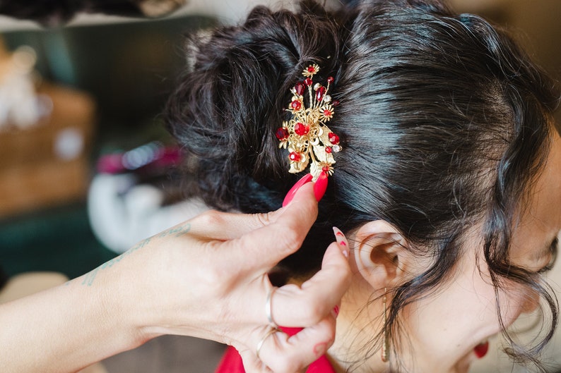 Peut inclure: Un accessoire pour cheveux dor&eacute; avec des pierres pr&eacute;cieuses rouges et des d&eacute;tails floraux est plac&eacute; dans des cheveux fonc&eacute;s. La pi&egrave;ce orn&eacute;e est une pince &agrave; cheveux d&eacute;corative, probablement pour une occasion sp&eacute;ciale, ajoutant une touche d'&eacute;l&eacute;gance.