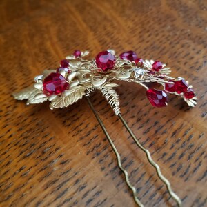 Peut inclure: Une &eacute;pingle &agrave; cheveux dor&eacute;e avec un motif floral, orn&eacute;e de pierres pr&eacute;cieuses rouges facett&eacute;es et de petits cristaux transparents. L'&eacute;pingle comporte deux longues et fines broches pour la fixation dans les cheveux. Le design comprend des d&eacute;tails de feuilles et de volutes.