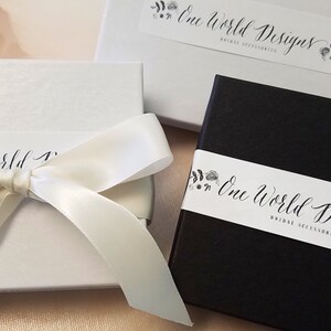 Peut inclure: Bo&icirc;tes cadeaux blanches et noires avec le texte "One World Designs Bridal Accessories". Une bo&icirc;te est blanche avec un ruban de satin nou&eacute; en n&oelig;ud. L'autre bo&icirc;te est noire avec une bande blanche.