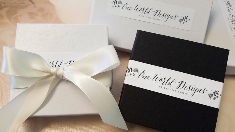 Peut inclure: Bo&icirc;tes cadeaux blanches et noires avec le texte "One World Designs Bridal Accessories". Une bo&icirc;te est blanche avec un ruban de satin nou&eacute; en n&oelig;ud. L'autre bo&icirc;te est noire avec une bande blanche.