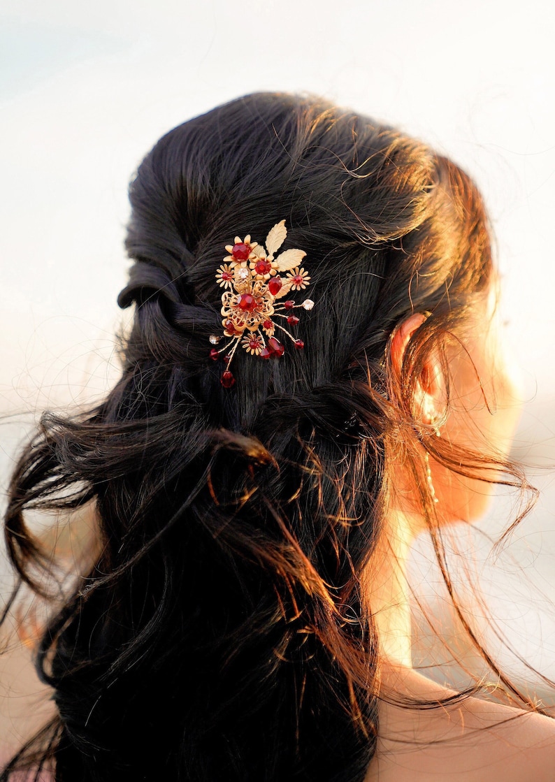 Peut inclure: Une barrette dor&eacute;e orn&eacute;e de pierres pr&eacute;cieuses rouges et transparentes et d'&eacute;l&eacute;ments floraux est fix&eacute;e dans des cheveux fonc&eacute;s. Cet accessoire de cheveux d&eacute;coratif est probablement destin&eacute; &agrave; une occasion sp&eacute;ciale, sur un fond flou.