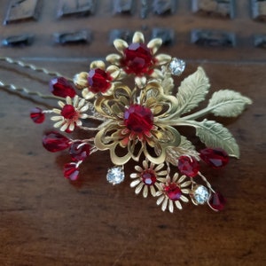 Peut inclure: &Eacute;pingle &agrave; cheveux dor&eacute;e avec un motif floral orn&eacute; de cristaux rouges et transparents. L'&eacute;pingle pr&eacute;sente une fleur centrale avec un grand cristal rouge, entour&eacute;e de petites fleurs et de gouttelettes de cristal. Les feuilles sont dor&eacute;es.