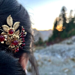 Peut inclure: Une barrette dor&eacute;e avec un motif floral et des pierres pr&eacute;cieuses rouges. La barrette pr&eacute;sente des feuilles et des fleurs d&eacute;taill&eacute;es, les pierres rouges ajoutant une touche de couleur. L'accessoire est plac&eacute; sur un arri&egrave;re-plan ext&eacute;rieur flou.