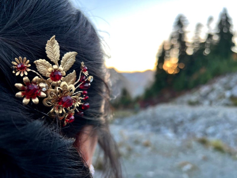 Peut inclure: Une barrette dor&eacute;e avec un motif floral et des pierres pr&eacute;cieuses rouges. La barrette pr&eacute;sente des feuilles et des fleurs d&eacute;taill&eacute;es, les pierres rouges ajoutant une touche de couleur. L'accessoire est plac&eacute; sur un arri&egrave;re-plan ext&eacute;rieur flou.