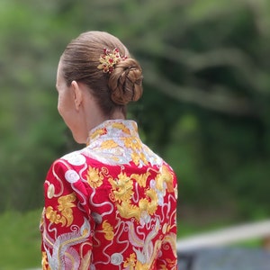 Peut inclure: Coiffure en chignon orn&eacute;e d'une barrette dor&eacute;e et rouge. La femme porte une veste rouge avec des broderies dor&eacute;es et argent&eacute;es repr&eacute;sentant des motifs de dragon et de ph&eacute;nix, un design traditionnel.