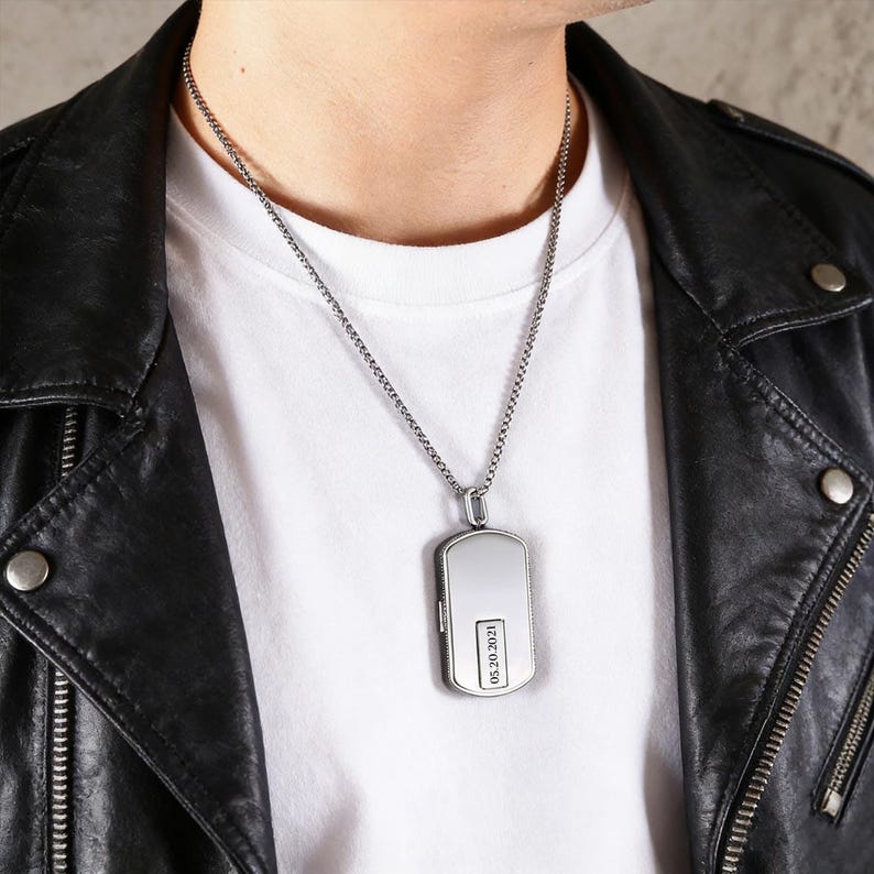 May include: A silver dog tag pendant necklace with a rectangular design. The pendant has a polished finish and a small rectangular section with the date "05.20.2021" engraved. The necklace is worn over a white t-shirt and a black leather jacket.