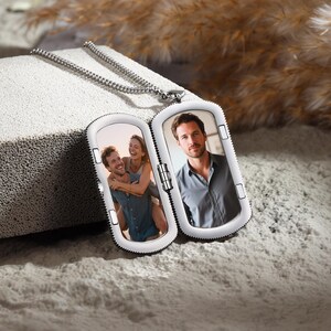 May include: A silver-colored locket necklace with a rectangular pendant. The pendant opens to reveal two photographs. One photo shows a couple, and the other shows a man. The locket is on a textured, light gray surface.