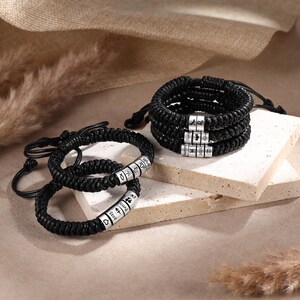 Könnte beinhalten: Mehrere schwarze geflochtene Armbänder mit silberfarbenen zylindrischen Perlen. Die Perlen haben eingravierte Texte und Symbole. Die Armbänder werden auf einer hellen, strukturierten Oberfläche mit neutralem Hintergrund präsentiert.