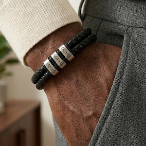 May include: Black braided leather bracelet with three silver name beads. The beads are engraved with the names "JAMES", "SOPHIA", and "OLIVER". The bracelet is worn on a person's wrist, with a neutral background.