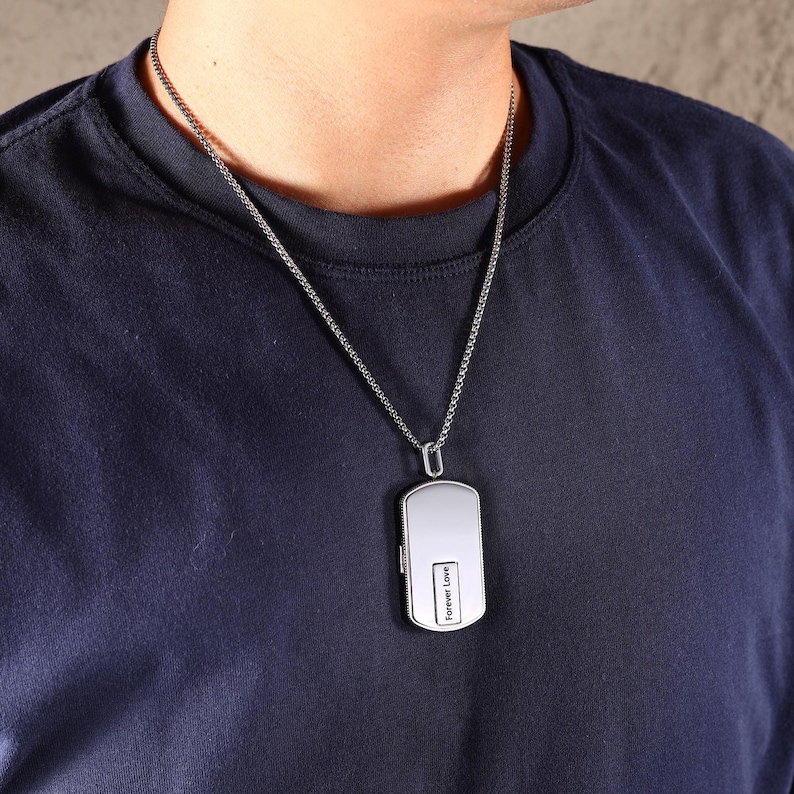 May include: Silver dog tag pendant necklace with the words "Forever Love" engraved on the front. The pendant hangs from a delicate chain. The person in the image is wearing a navy blue t-shirt.