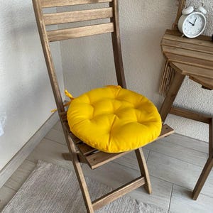 Puede incluir: Un coj&iacute;n de asiento redondo, acolchado y de color amarillo brillante con lazos de tela, colocado sobre una silla plegable de madera. El coj&iacute;n presenta un patr&oacute;n de costura circular y se sit&uacute;a sobre el asiento de listones de la silla.