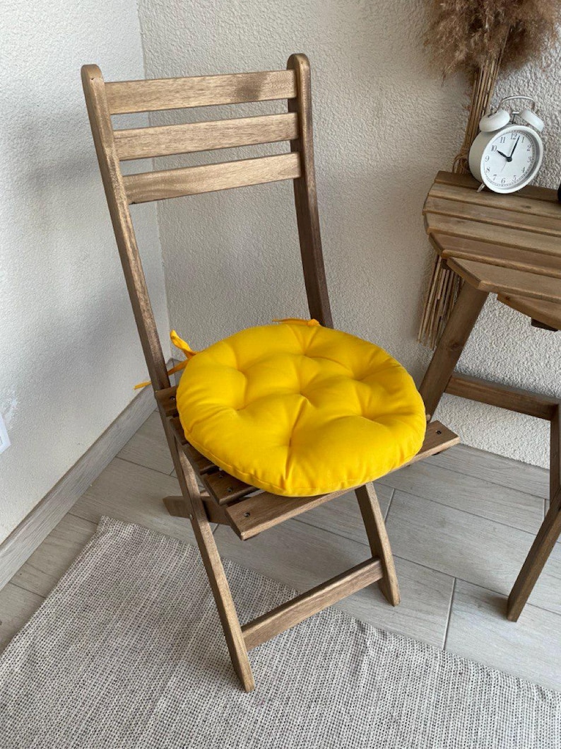 Puede incluir: Un coj&iacute;n de asiento redondo, acolchado y de color amarillo brillante con lazos de tela, colocado sobre una silla plegable de madera. El coj&iacute;n presenta un patr&oacute;n de costura circular y se sit&uacute;a sobre el asiento de listones de la silla.