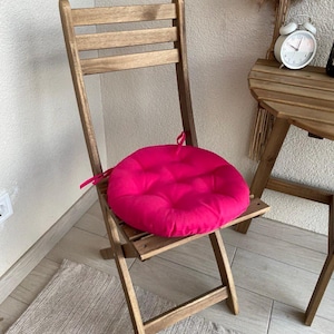 Puede incluir: Una silla plegable de madera con un coj&iacute;n de asiento circular de color fucsia brillante. El coj&iacute;n est&aacute; acolchado y cuenta con finas cintas de tela en los laterales para asegurarlo al marco de la silla. La silla tiene un respaldo de listones.