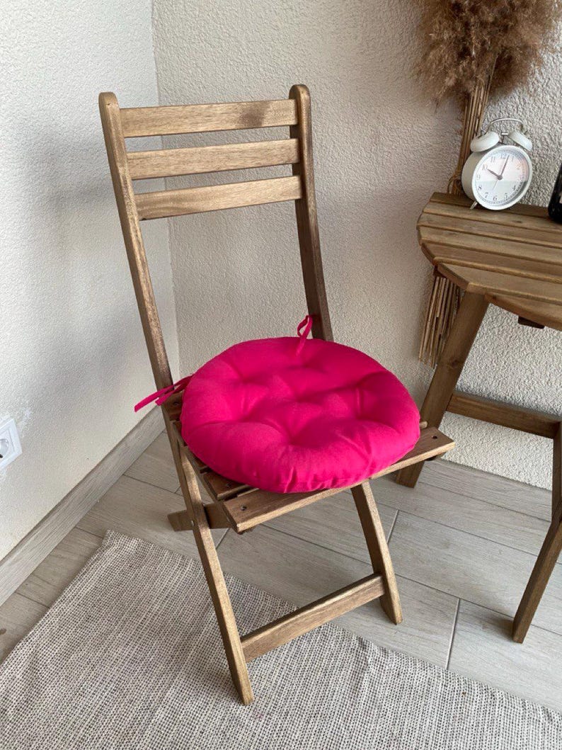 Puede incluir: Una silla plegable de madera con un coj&iacute;n de asiento circular de color fucsia brillante. El coj&iacute;n est&aacute; acolchado y cuenta con finas cintas de tela en los laterales para asegurarlo al marco de la silla. La silla tiene un respaldo de listones.