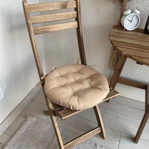 Puede incluir: Un coj&iacute;n de asiento redondo y beige con capiton&eacute; y lazos de tela en los laterales, colocado sobre una silla plegable de madera. El coj&iacute;n presenta un bot&oacute;n central y una estructura acolchada dise&ntilde;ada para ajustarse al asiento de la silla.