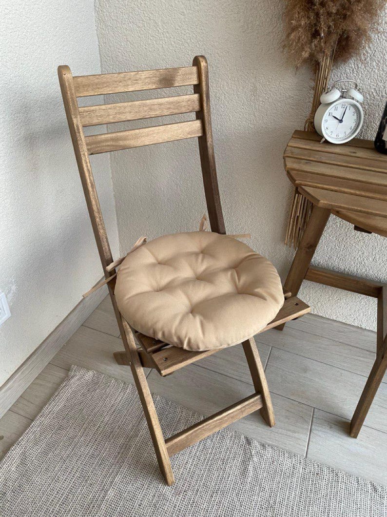 Puede incluir: Un coj&iacute;n de asiento redondo y beige con capiton&eacute; y lazos de tela en los laterales, colocado sobre una silla plegable de madera. El coj&iacute;n presenta un bot&oacute;n central y una estructura acolchada dise&ntilde;ada para ajustarse al asiento de la silla.