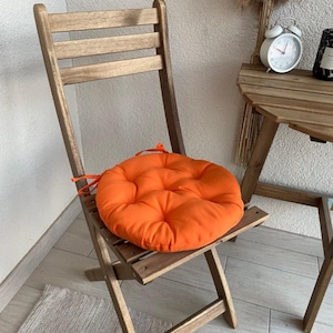Puede incluir: Un coj&iacute;n de asiento redondo, acolchado y de color naranja brillante con lazos de tela, colocado sobre una silla plegable de madera. El coj&iacute;n presenta un patr&oacute;n de costuras circulares y una textura suave, creando un contraste vibrante.