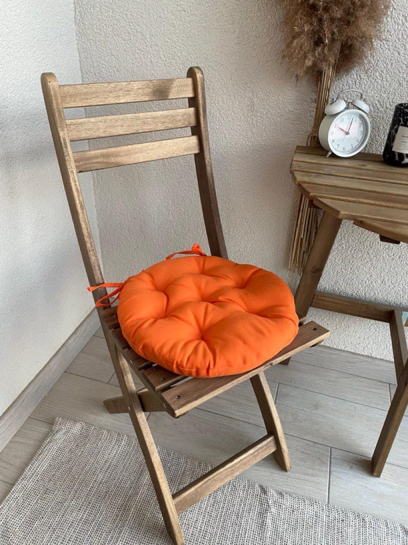 Puede incluir: Un coj&iacute;n de asiento redondo, acolchado y de color naranja brillante con lazos de tela, colocado sobre una silla plegable de madera. El coj&iacute;n presenta un patr&oacute;n de costuras circulares y una textura suave, creando un contraste vibrante.
