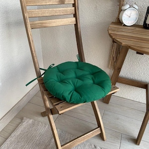 Puede incluir: Un coj&iacute;n de asiento redondo y capiton&eacute; en color verde bosque con lazos de tela en los laterales, colocado sobre una silla plegable de madera. El coj&iacute;n presenta un patr&oacute;n de costuras circulares y est&aacute; hecho de tela lisa y suave.