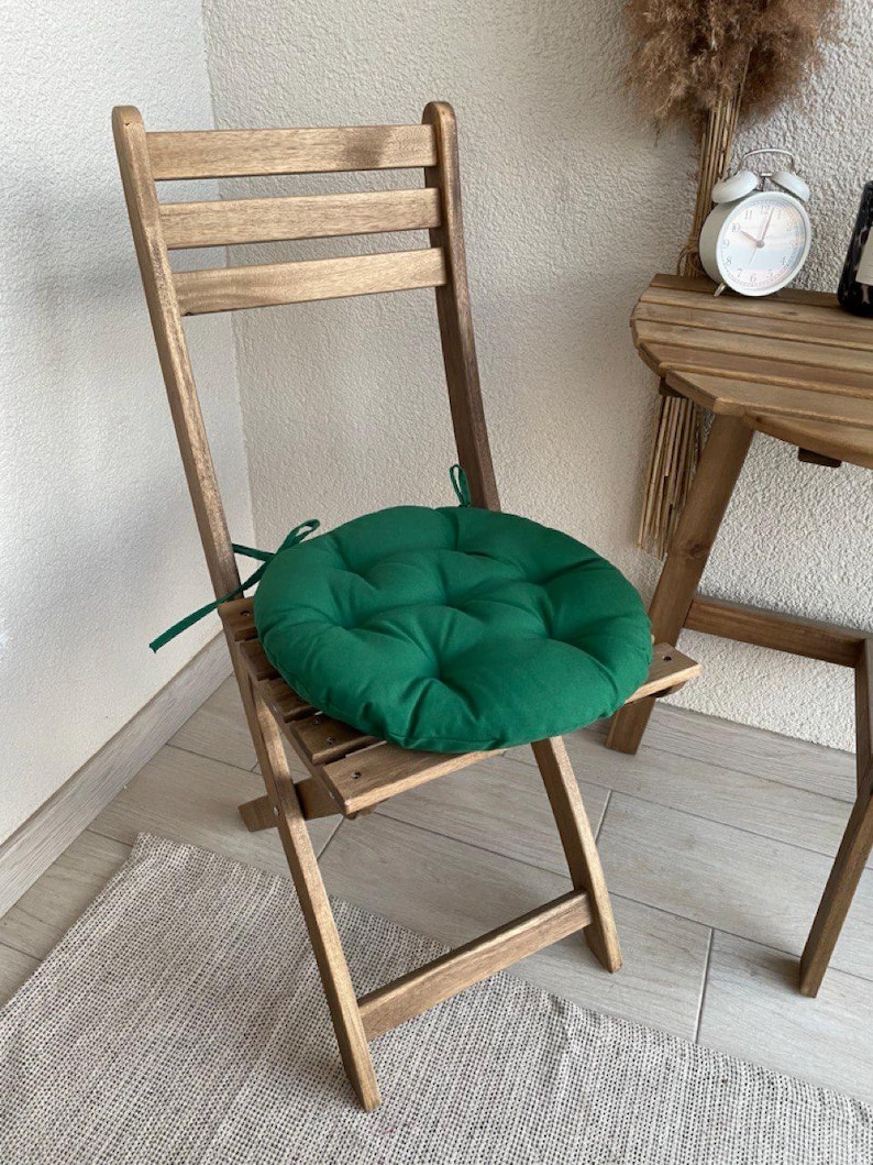 Puede incluir: Un coj&iacute;n de asiento redondo y capiton&eacute; en color verde bosque con lazos de tela en los laterales, colocado sobre una silla plegable de madera. El coj&iacute;n presenta un patr&oacute;n de costuras circulares y est&aacute; hecho de tela lisa y suave.