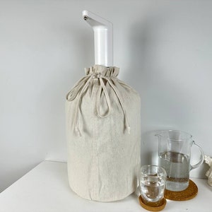 May include: A white water dispenser with a spout and a white linen cover. The dispenser is sitting on a white table with a glass jug and two glasses of water.