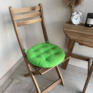 Puede incluir: Un coj&iacute;n de asiento circular de color verde brillante, acolchado y con lazos de tela en los laterales, colocado sobre una silla plegable de madera marr&oacute;n. El coj&iacute;n presenta una textura suave y mullida con hendiduras.