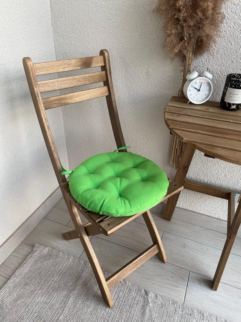 Puede incluir: Un coj&iacute;n de asiento circular de color verde brillante, acolchado y con lazos de tela en los laterales, colocado sobre una silla plegable de madera marr&oacute;n. El coj&iacute;n presenta una textura suave y mullida con hendiduras.