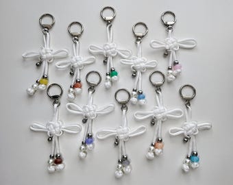 May include: A collection of white knotted keychains with silver-tone hardware. Each keychain features a cluster of colorful beads in various shades, including yellow, orange, green, blue, and pink. The keychains are arranged on a white surface.