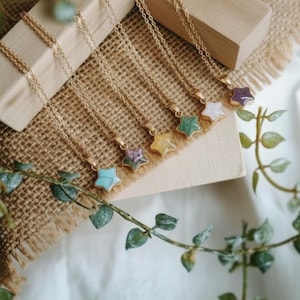 Peut inclure: Plusieurs colliers dorés avec des pendentifs en forme d'étoile de différentes couleurs. Les pendentifs sont en pierres précieuses. Les colliers sont présentés sur un bloc de bois et du tissu de jute, avec des feuilles vertes au premier plan.