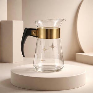 May include: Clear glass pitcher with a gold band and black handle. The pitcher has a flared top and is decorated with gold starburst designs. The pitcher is sitting on a white pedestal.