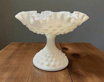 Può includere: Una ciotola a piedistallo in vetro latte bianco, stile hobnail, con bordo arricciato. La ciotola poggia su un gambo corto e una base rotonda. Il motivo hobnail ricopre la base e il fondo della ciotola.