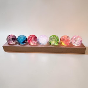 May include: Seven colorful glass spheres, each with a unique internal design, are arranged on a rectangular wooden base. The spheres display a range of colors, including pink, blue, red, and green, creating a vibrant display. The wooden base is a light brown color.