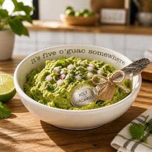 May include: A white ceramic bowl filled with green guacamole topped with diced red onion and cilantro. The rim features the black text "It's five o'guac somewhere." A silver spoon with the text "HOLY GUACAMOLE" rests inside, tied with twine.