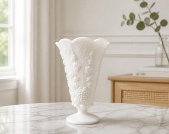 Può includere: Un vaso in vetro opalino bianco con bordo smerlato e un motivo in rilievo di uva e foglie. Il vaso ha un corpo affusolato che si allarga verso l'alto e poggia su una base circolare, caratterizzato da una finitura liscia e opaca.