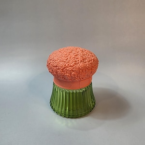May include: A decorative glass jar with a coral-colored, floral-textured lid. The jar has a green, ribbed base and a coral-colored band. The jar is a unique decorative item.