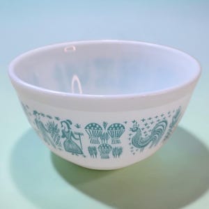 May include: White mixing bowl with a teal design featuring a rooster, woman, and other farm-themed illustrations. The bowl has a rounded shape and a smooth, glossy finish. A vintage kitchenware item.