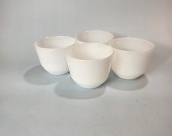 May include: Four white ceramic bowls are arranged on a light blue surface. The bowls have a rounded shape with a wide opening and a smooth, glossy finish. The bowls are evenly spaced and appear to be empty.