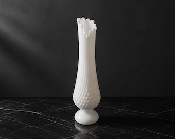 Può includere: Un vaso alto in vetro opalino bianco caratterizzato da un motivo a rilievo sulla parte inferiore bombata, un collo sottile e un bordo smerlato. Il vaso poggia su una base circolare a piedistallo, mostrando una classica struttura in vetro opaco.