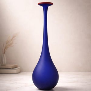 May include: A tall, elegant blue vase with a teardrop shape and a narrow neck. The vase has a matte finish and a small, red circular top. The vase is displayed on a white surface, suggesting a decorative home accent.
