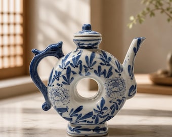 Può includere: Una teiera in ceramica bianca con un corpo circolare a forma di ciambella con un foro centrale. Il pezzo è decorato con motivi floreali e di foglie dipinti in blu. Include un piccolo coperchio, un beccuccio curvo e un manico a forma di animale.