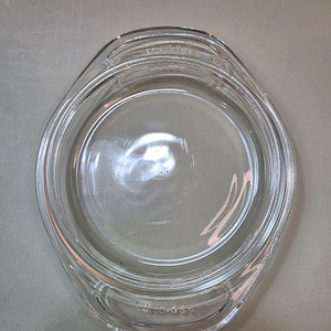 May include: Clear glass Pyrex baking dish with a round interior and an oval-shaped exterior. The dish has two handles and is marked with text near the handles. The dish is empty and sits on a neutral surface.