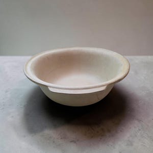 May include: A light beige, round bowl with a wide rim and two small handles. The bowl has a simple, minimalist design and appears to be made of a natural material. The bowl is empty and sits on a gray surface.