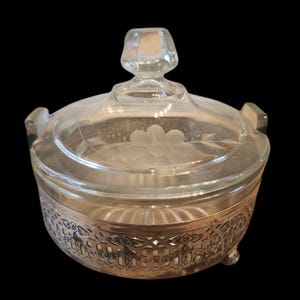 May include: A clear glass jar with a silver-toned metal base and lid. The jar has a floral design etched into the glass and the base has a decorative, openwork design.